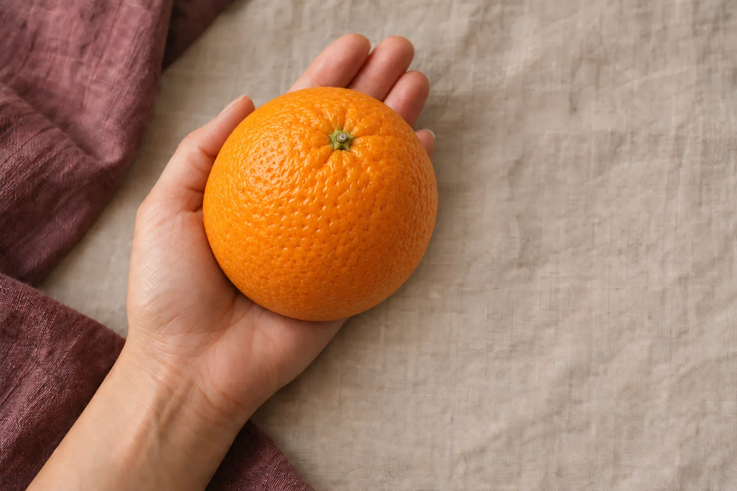 Eine einzelne Orange liegt in einer offenen Handfläche auf beigem Leinen