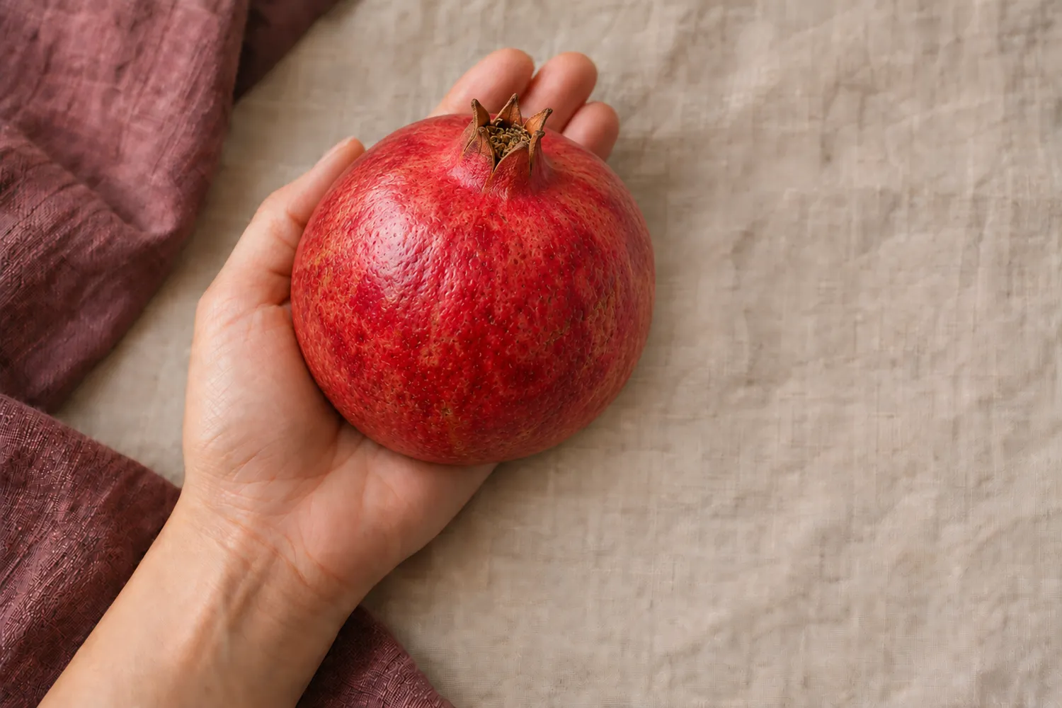 Ein einzelner roter Granatapfel liegt in einer offenen Handfläche auf beigem Leinen