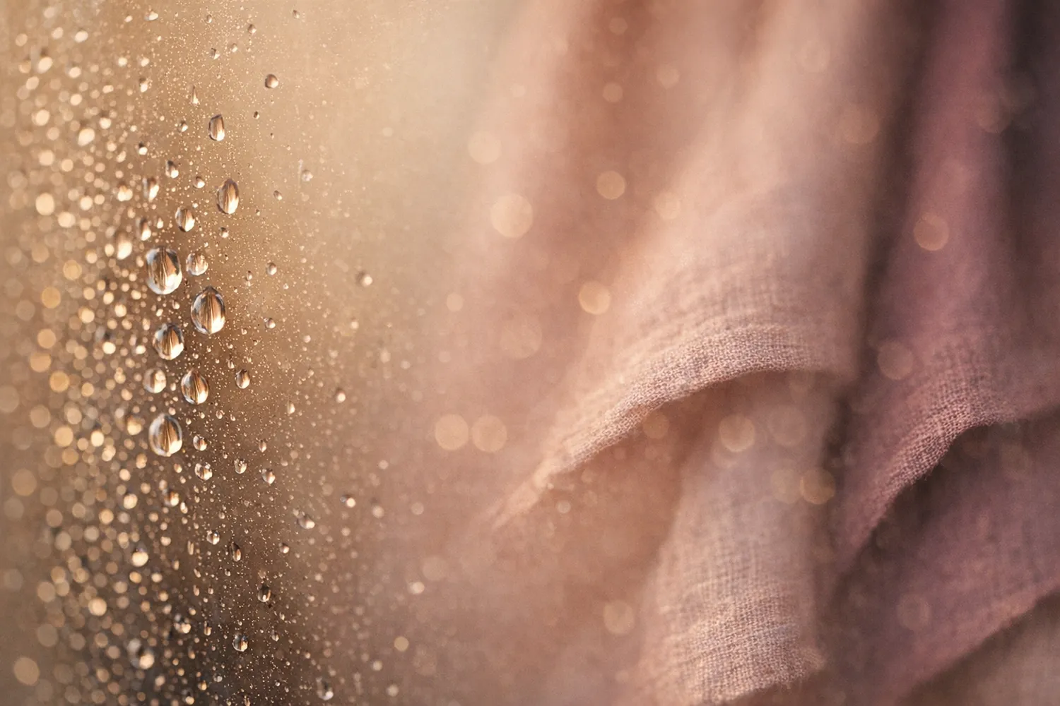 Gocce d’acqua su una lastra di vetro nella calda luce del mattino, davanti a un tessuto di lino rosé
